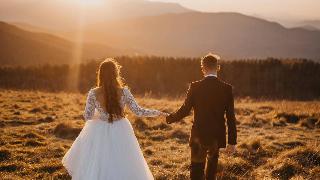 A man and woman walking hand in hand in a filed, wearing a wedding dress and suit.