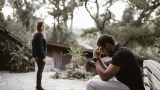 A man sits on a bench in thought while a woman looks at him in the midground.