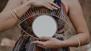 A woman in a pattered dress holding an eye-shaped mirror in front of her chest.