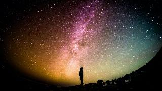 A silhouette of a man standing below a brilliant sky of rainbow-tinted stars.