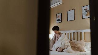 A woman sitting on her bed, wrapped in a comforter, as seen through her doorway.