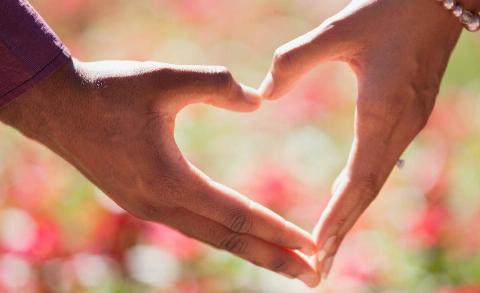 Two hands making a heart shape together against a blurred pink and green backdrop.