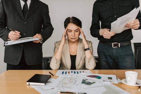 A woman seated at a desk with her hands to her temples, two male coworkers standing on either side of her, the desk is covered in papers.