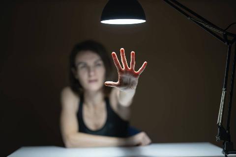 A woman seated at a table lit with an overhead lamp, reaching out towards the camera.