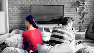 A greyscale image of a couple sitting on a couch in their living room, turned away from one another, but the woman is still in color and glowing blew.