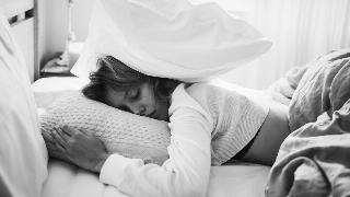 A greyscale image of a woman laying in bed, a pillow on top of her head.