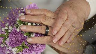 Elderly couple holding hands and showing their wedding rings and bouquet, constellations decorating the corners of the image.