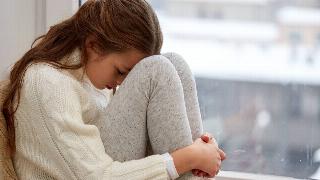 A young girl seated on a windowsill, her knees to her chest, forehead resting on her knees.