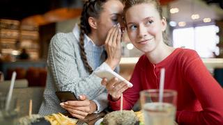 Two casual friendly girls with smartphones gossipping by served table in cafe.