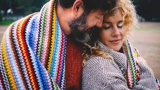 Romantic adult couple hugging while covered by a woolen colorful blanket at the outdoor park.