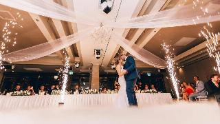 A low-angle shot of a couple kissing at a wedding, sparklers flying around them and a set table full of friends behind them.