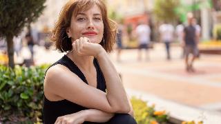 An elegant woman sitting outside, looking at the camera, resting her chin on her hand.