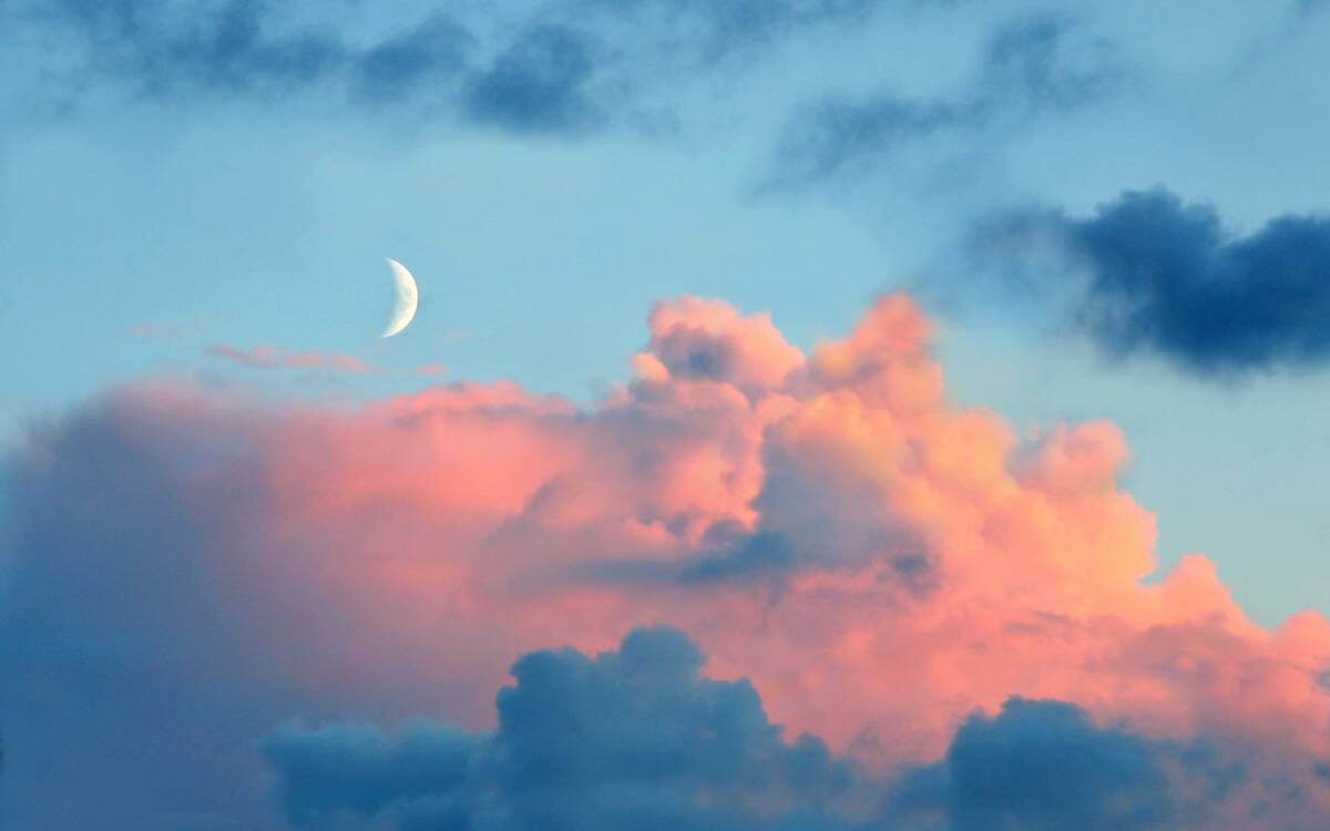 A new moon in a bright blue sky with colorful pink and blue clouds.