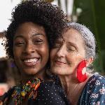 Two women embracing amicably, pressing their heads together, one behind the other, both smiling.