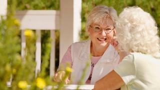 Two older women sitting at a table outside talking with one another, smiling.