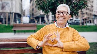 A woman in an orange sweater on a park bench, making a heart with her fingers in front of her chest.