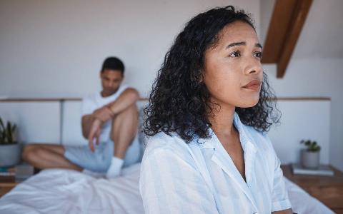 A woman sitting on her bed, looking pained, a man sitting in the background looking regretful.