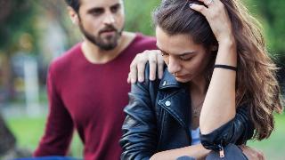 A couple fighting, the woman with a hand in her hair as she looks sad while the man insistently puts a hand on her shoulder.