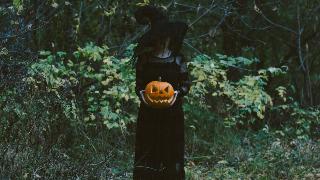 A woman in a witch outfit standing outside in the greenery, holding a jack-o-lantern.