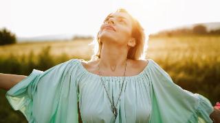 A woman in a seafoam green dress, standing outside, looking up and smiling as the sun shines on her face.