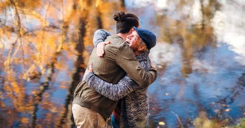 A father and son hugging by the water that's reflecting an autumn scene around them.