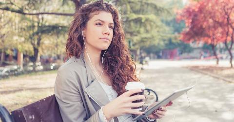 A woman sitting on a park bench, earbuds in and connected to an iPad, a coffee cup in her hand.