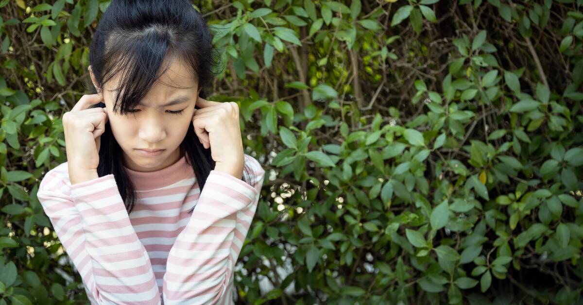 A girl looking annoyed as she plugs her ears with her fingers.