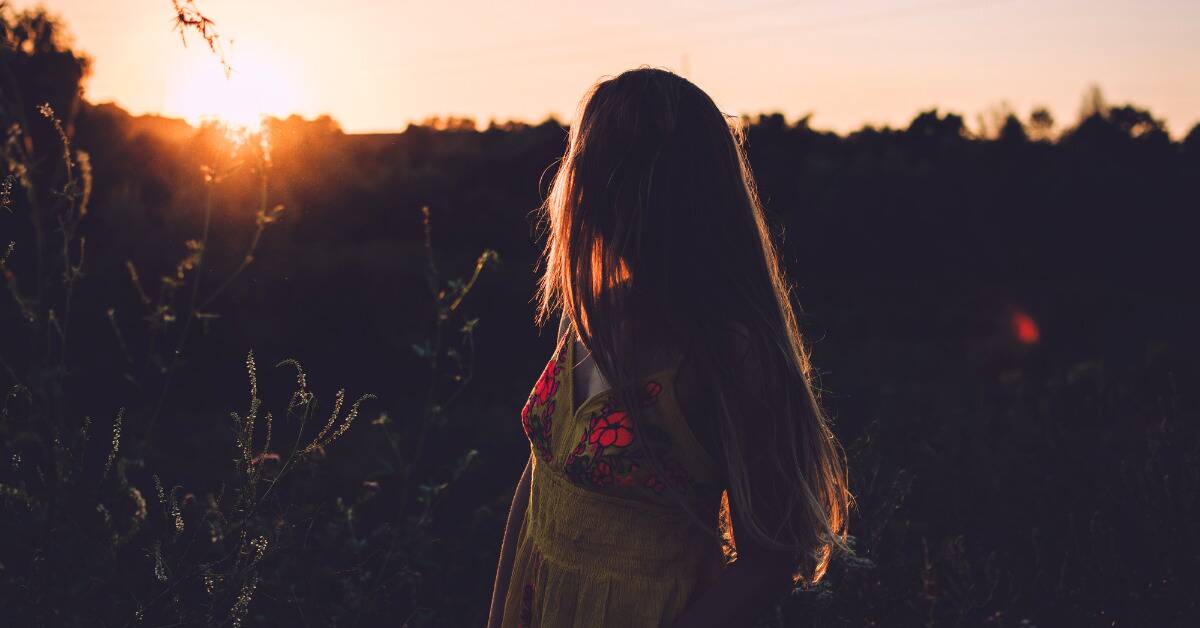 A woman looking over her shoulder at the setting sun while standing outside.