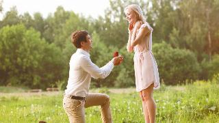 A young couple in a field, the man on one knee proposing to the woman, both smiling and looking excited.