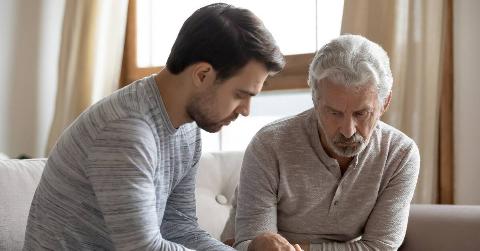 A father and his adult son sitting on a couch, both looking downward in a dejected manner.