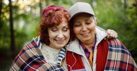 Two friends standing shoulder to shoulder as they walk outside, a blanket wrapped around them both, each with an earbud in listening to something on one of their phones.