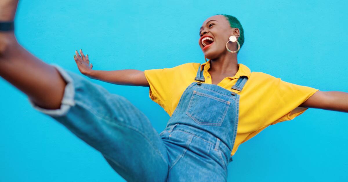 A woman in a bright yellow shirt with green hair smiling and kicking her leg up against a bright blue background.