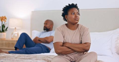 A woman sitting on a bed with her arms crossed, seeming frustrated. Behind her is a man with his arms crossed, looking away from her.