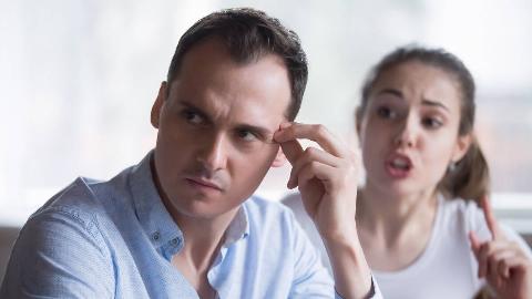 A man with his fingers to his temple, looking annoyed as a woman speaks fervently behind him.