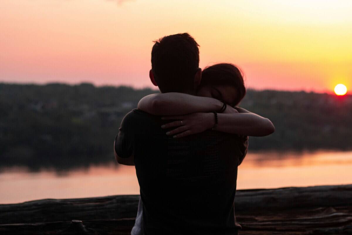 A man and a woman hug by the water with the sun setting in the background.