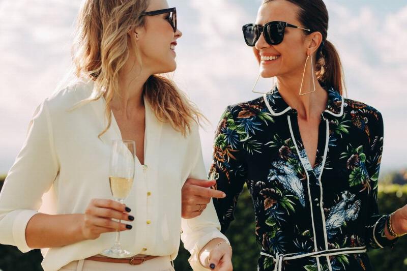 Two stylish friends smiling as they walk and chat outside, arms linked, holding glasses of champagne.