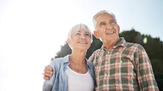 A couple standing outside together, shot from a low angle against a bright sky, the man with his arm around the woman, both smiling as they look into the distance.