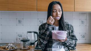 A woman standing in her kitchen, eating a spoonful of ice cream from a tub.