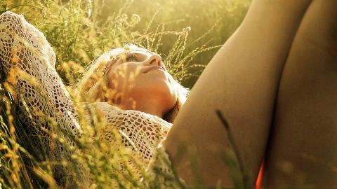 A close, low shot of a woman laying in the grass, hands behind her head, looking to the sky as the sun shines on her.