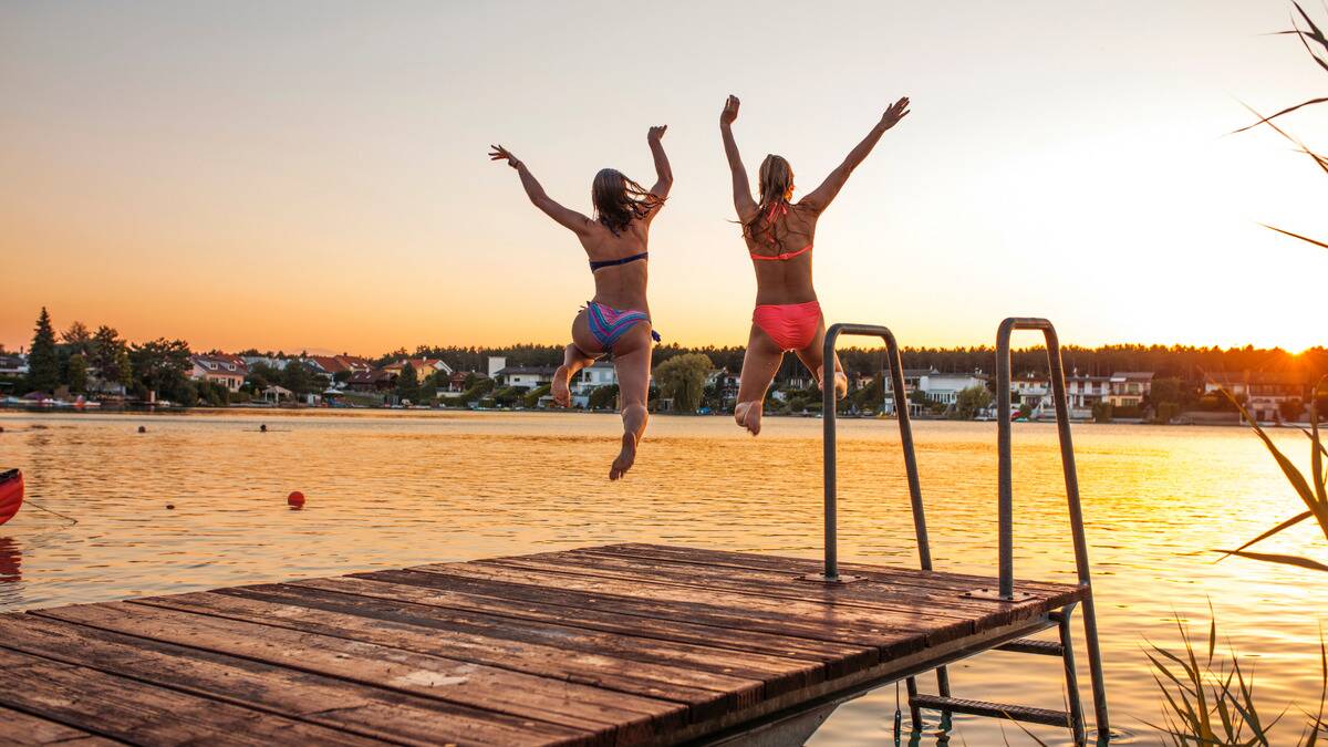 Two woman in bathing suits jumping off a short dock into the water during a sunset.