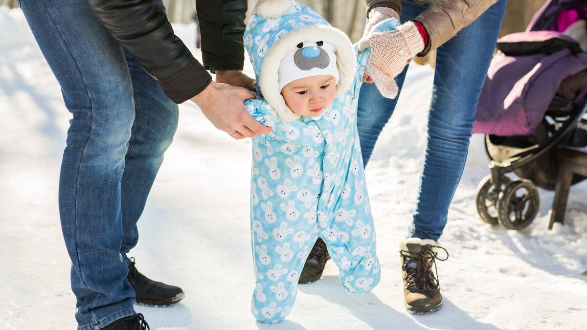 A baby in a onesie whose parents are helping it walk over the snow.