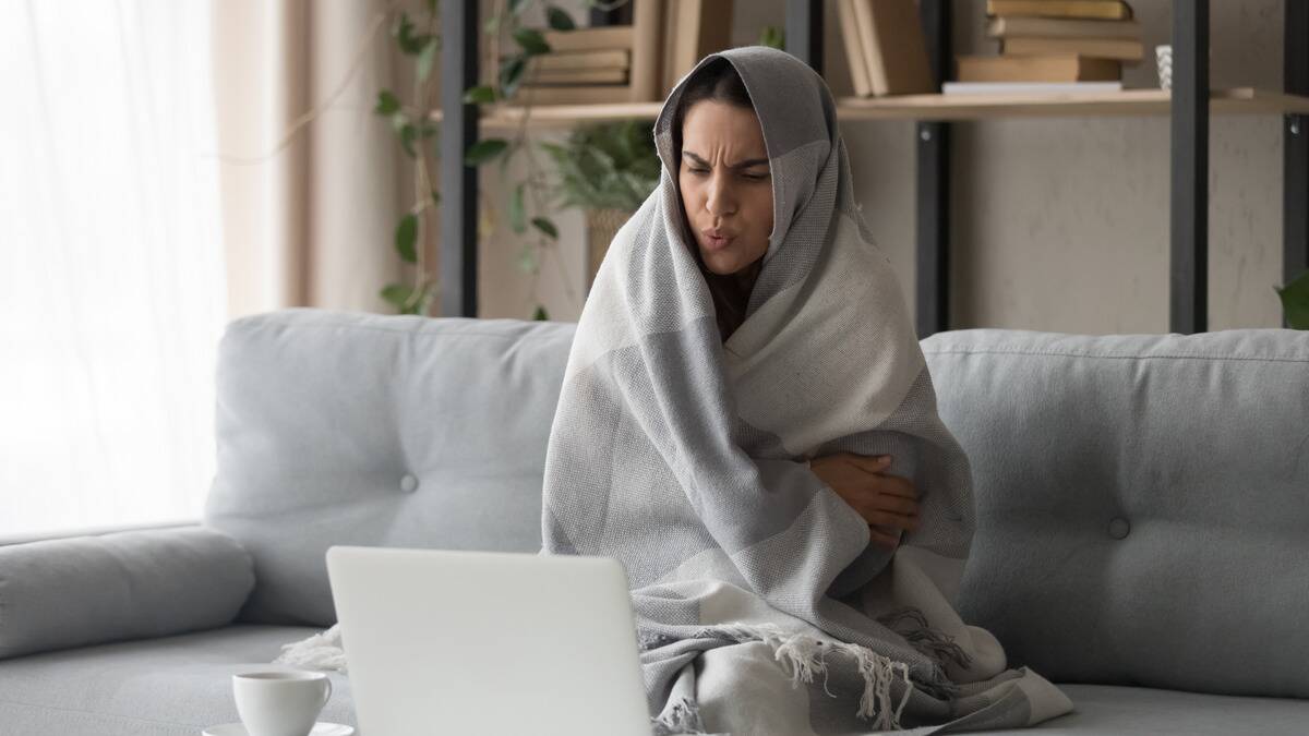 A woman wrapped up in a blanket while sitting on her couch, looking cold, watching something on her laptop.