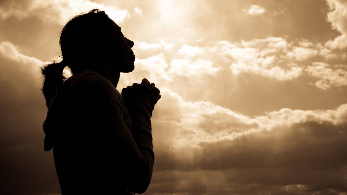 A woman with her hands together in prayer or pleading, silhouetted against a sunrise sky.