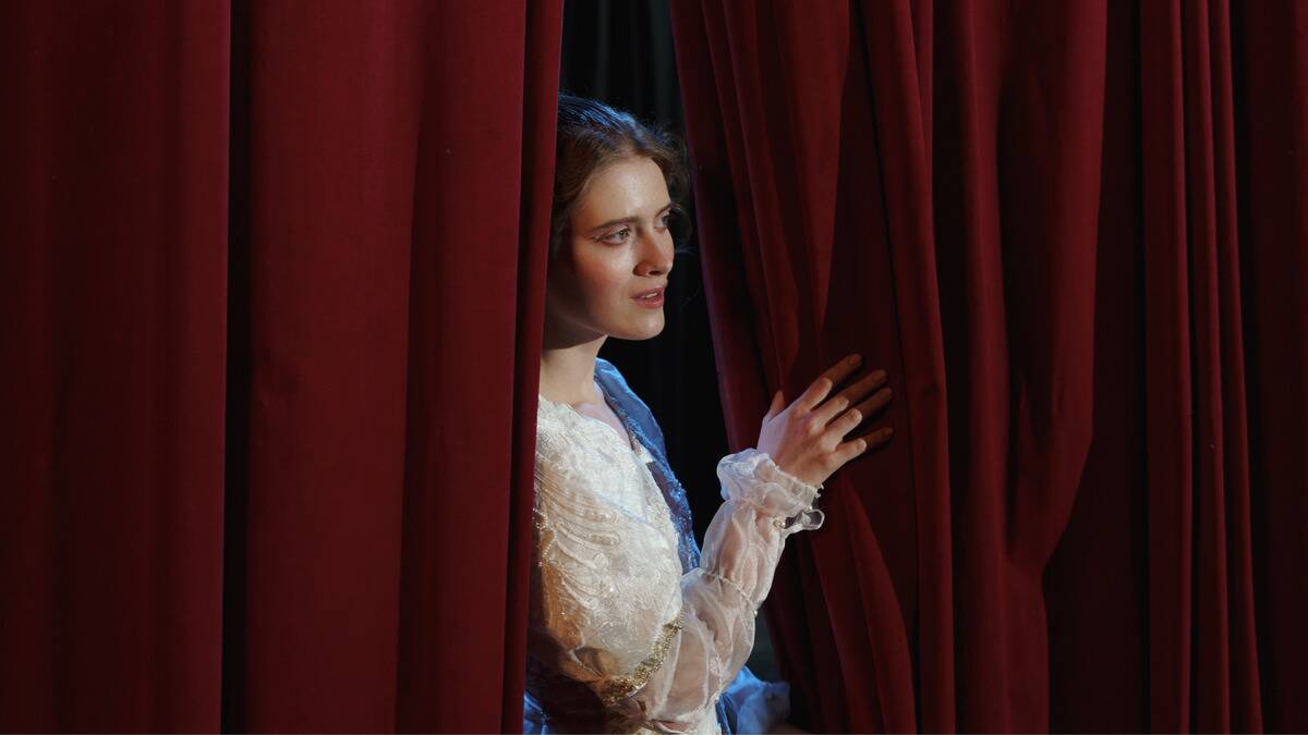 A woman in a play peering through the curtain, in a white tulle dress.