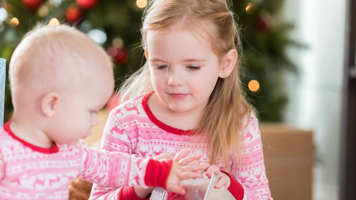 An older sister with her baby sibling in matching Christmas pajamas, the baby reaching out for the gift box she's holding.