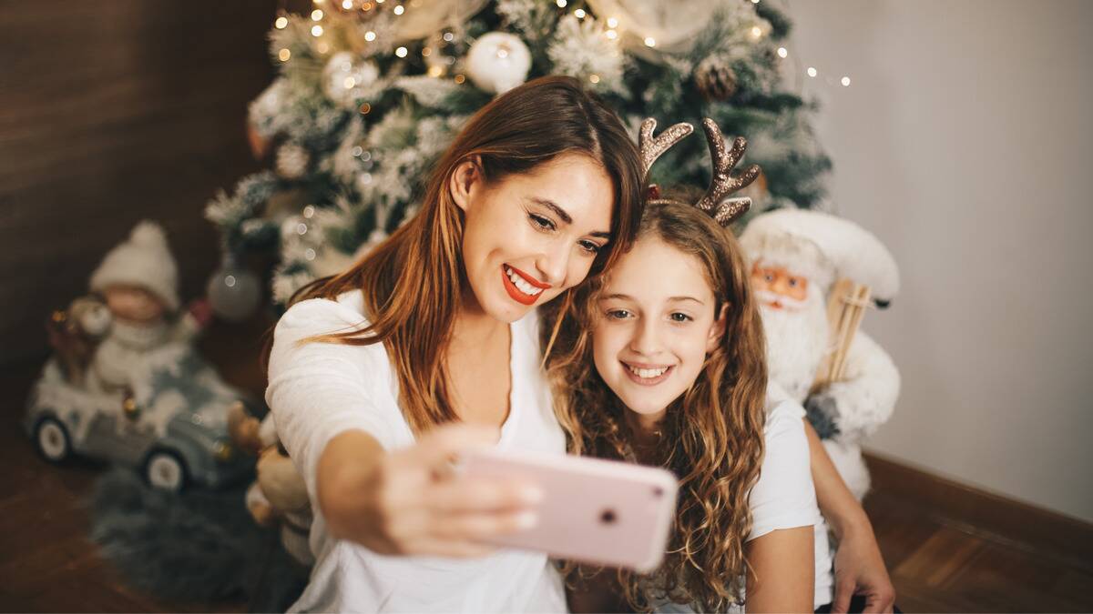 An older sister taking a selfie with her younger sister in front of a Christmas tree. 