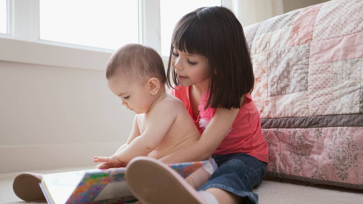 A young girl with her younger sibling sitting between her legs, both looking down a large, open storybook.