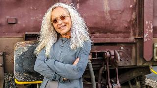 An older woman standing confidently with crossed arms in front of what appears to be an industrial truck or train car.