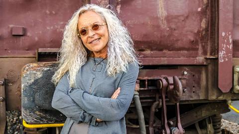 An older woman standing confidently with crossed arms in front of what appears to be an industrial truck or train car.