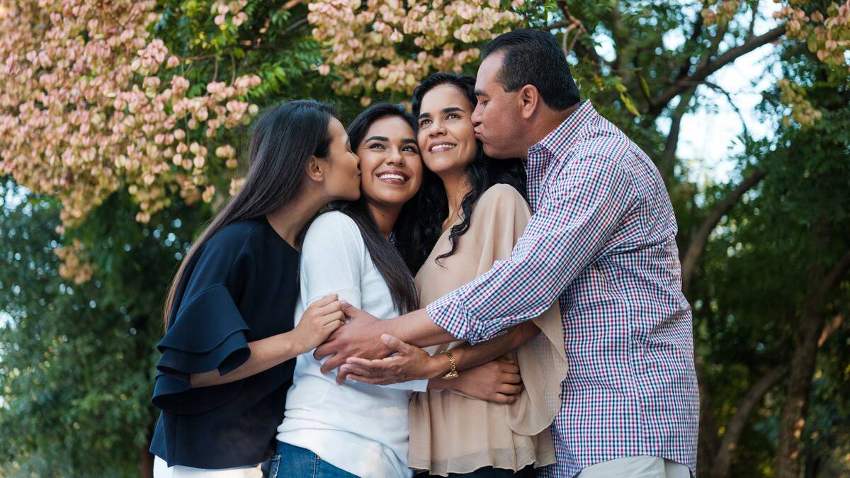 A family posing outside together. The father is kissing his wife on the cheek, one sister is kissing the other sister on the cheek, and they're all embracing each other in a line.